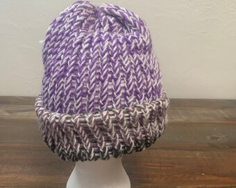 May include: Purple and white knitted beanie with a white pom pom on top. The beanie has a chevron pattern and a folded brim.