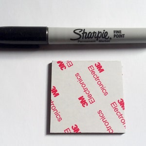 Pode incluir: Um marcador permanente preto Sharpie Fine Point posicionado acima de uma almofada adesiva quadrada. O verso branco da almofada apresenta um padr&atilde;o vermelho repetitivo com o texto 3M Electronics, indicando a marca e o uso do item.