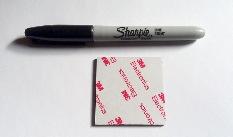 Pode incluir: Um marcador permanente preto Sharpie Fine Point posicionado acima de uma almofada adesiva quadrada. O verso branco da almofada apresenta um padr&atilde;o vermelho repetitivo com o texto 3M Electronics, indicando a marca e o uso do item.