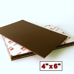 Pode incluir: Uma pilha de folhas retangulares de espuma castanha com verso adesivo, cada uma medindo 10,16 cm por 15,24 cm. O verso adesivo branco apresenta o logótipo vermelho da 3M Electronics impresso repetidamente na superfície do material.