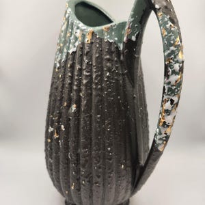 May include: A black and green ceramic pitcher with a textured surface and gold accents. The pitcher has a wide mouth and a handle.