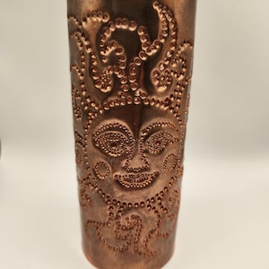 Puede incluir: Una linterna de cobre para velas con un diseño de cara decorativo. La linterna tiene una superficie texturizada con pequeños puntos y una parte superior festoneada.