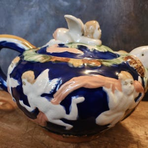 Puede incluir: Una tetera de cerámica azul y blanca con un pico de delfín y figuras de querubines. La tetera está decorada con diseños florales y de volutas.