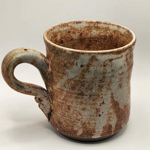 Puede incluir: Una taza de cerámica hecha a mano con un aspecto rústico y texturizado. La taza presenta un cuerpo redondeado y un asa robusta. La paleta de colores incluye tonos marrones, grises y toques de azul, creando una estética natural y terrosa.