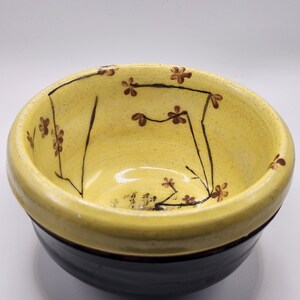 May include: A small yellow ceramic bowl with a black rim and brown floral design.