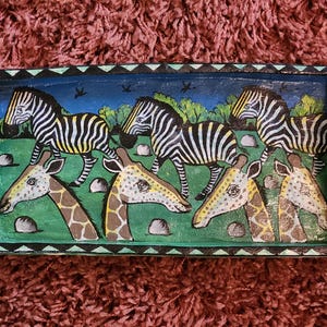 Puede incluir: Una bandeja de servir de madera con fondo azul y un diseño pintado de colores de jirafas y cebras. La bandeja tiene un borde decorativo con un patrón ondulado.