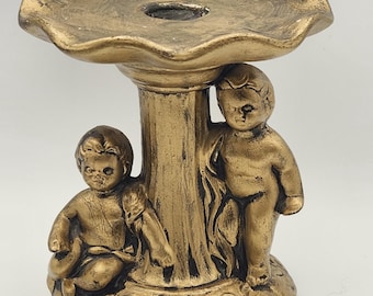 May include: A gold-colored cherub candle holder. The candle holder features a fluted top with a central hole, supported by a column. Two cherub figures are sculpted on the base, surrounded by leaf detailing.