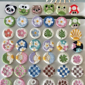 Op de afbeelding: Een verzameling kleurrijke, ronde, getufte onderzetters. Ontwerpen omvatten een panda, kikker, beer, bloemen, bladeren, paddenstoelen en geometrische patronen. De onderzetters zijn gerangschikt op een wit oppervlak.