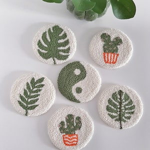 Op de afbeelding: Zes ronde, witte onderzetters met groene en oranje borduurwerkontwerpen. De ontwerpen omvatten een monstera-blad, een varen, een cactus, een yin-yang-symbool en twee verschillende cactusontwerpen.