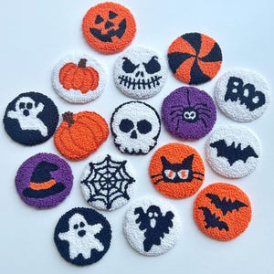 Pode incluir: Uma coleção de porta-copos circulares feitos com punch needle com designs de Halloween, incluindo abóboras, fantasmas, caveiras, morcegos, uma aranha, uma teia, um chapéu de bruxa, um gato preto e a palavra BOO em texturas de fios laranja, preto, branco e roxo.