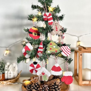 May include: A small Christmas tree decorated with handmade ornaments. The ornaments include a Santa belt, gingerbread man, candy cane, mittens, and a house. The ornaments are red, white, green, and tan. The tree sits on a wooden tray with pine cones.