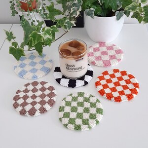 May include: Six round crocheted coasters with a checkered pattern in various colors, including blue, pink, brown, green, and orange. The coasters are arranged on a white surface with a glass of iced coffee in the center. The glass has a black and white checkered coaster underneath it. The glass has the text "Good Morning" on it.