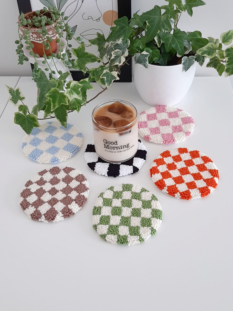 May include: Six round crocheted coasters with a checkered pattern in various colors, including blue, pink, brown, green, and orange. The coasters are arranged on a white surface with a glass of iced coffee in the center. The glass has a black and white checkered coaster underneath it. The glass has the text "Good Morning" on it.