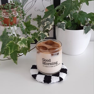 May include: A glass of iced coffee with ice cubes in a clear glass with the text "Good Morning It's time to wake up and shine" on the side, sitting on a black and white checkered coaster.