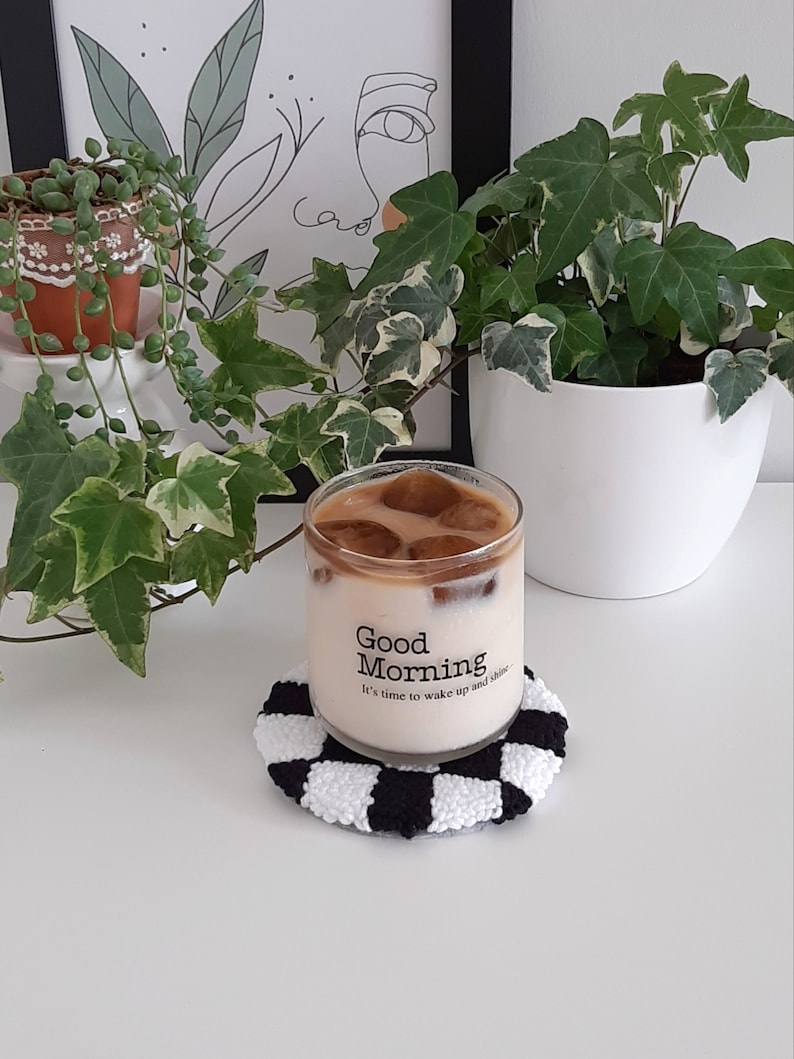 May include: A glass of iced coffee with ice cubes in a clear glass with the text "Good Morning It's time to wake up and shine" on the side, sitting on a black and white checkered coaster.