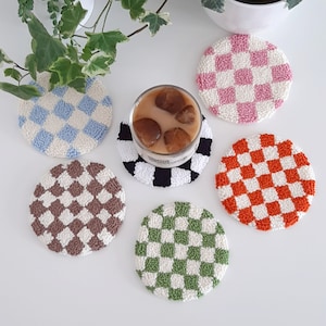 May include: Six round crocheted coasters with a checkered pattern in various colors, including pink, orange, brown, green, and blue. The coasters are on a white surface with a glass of iced coffee.