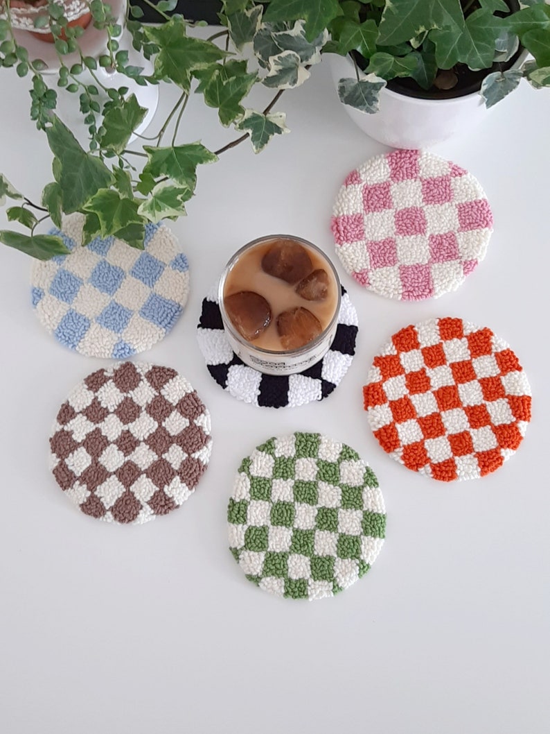 May include: Six round crocheted coasters with a checkered pattern in various colors, including pink, orange, brown, green, and blue. The coasters are on a white surface with a glass of iced coffee.