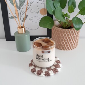 May include: A glass of iced coffee with ice cubes in a clear glass with the text "Good Morning It's time to wake up and shine." on the side. The glass is sitting on a brown and white crocheted coaster.