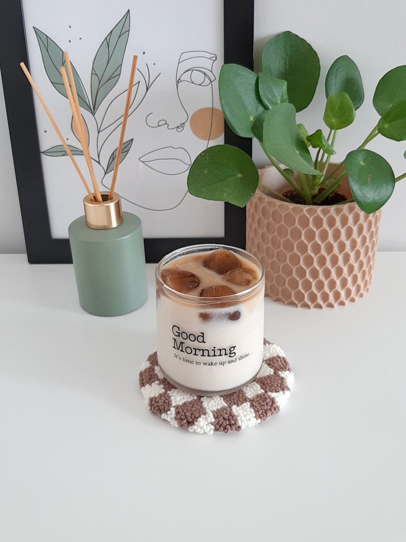 May include: A glass of iced coffee with ice cubes in a clear glass with the text "Good Morning It's time to wake up and shine." on the side. The glass is sitting on a brown and white crocheted coaster.