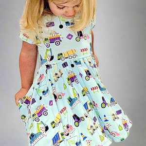 May include: A young girl wearing a light blue dress with a construction vehicle print. The dress has short sleeves and a gathered skirt. The girl is standing on a gray background.