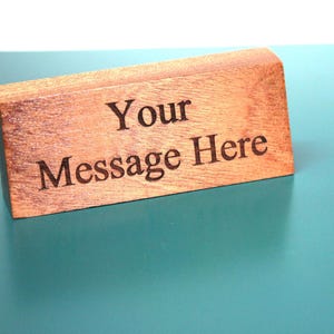 May include: A rectangular wooden sign with the engraved text "Your Message Here." The sign is made of light brown wood and sits on a teal surface. The text is in a simple, sans-serif font.