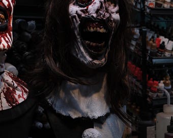 May include: A detailed bust of a pale, zombie-like figure with long dark hair, wearing a crown of thorns with red blood-like stains. The figure has an open mouth, one yellow eye, and a white collar. A black base displays the text DAMIEN LEONE'S TERRIFIER.