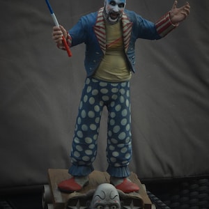 Può includere: Statuetta di un clown con trucco blu, una giacca blu a righe rosse e bianche e pantaloni blu a pois bianchi. Il clown è in piedi su una piattaforma di legno con una faccia grottesca.