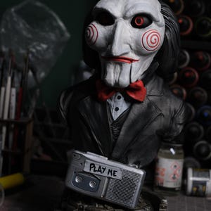 Puede incluir: Un busto de Billy the Puppet con cara blanca, mejillas en espiral rojas, ojos negros con iris rojos y una pajarita roja. La figura viste un traje negro y está colocada sobre un reproductor de casetes plateado con el texto PLAY ME.