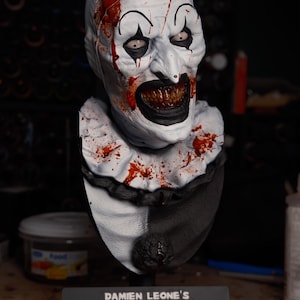 May include: A bust of a clown character with a white face, black eye makeup, and a wide mouth showing dark teeth. The clown wears a white ruffled collar with red blood splatters and a black and white outfit. A plaque reads DAMIEN LEONE'S TERRIFIER.
