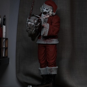 Könnte beinhalten: Figur einer Santa Claus-ähnlichen Figur mit Totenkopfgesicht, rotem Anzug mit weißem Besatz und Weihnachtsmütze. Die Figur hält eine blutbedeckte Kettensäge. Auf dem Sockel steht "Damien Leone's Terrifier 3".