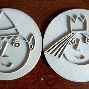 May include: Two 3D printed cookie cutters, one shaped like an elf and the other shaped like a princess. Both cutters are grey and have a detailed design.