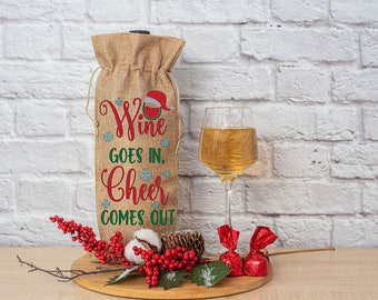 以下が含まれることがあります： 赤と緑で「Wine goes in, Cheer comes out」の文字が入ったジュートワインボトルバッグ。琥珀色の液体が入ったワイングラス、赤いキャンディー、装飾的なベリーが木の板の上に置かれています。サンタの帽子がバッグに付いています。