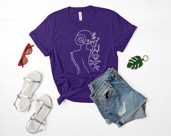 Peut inclure: T-shirt à manches courtes de couleur violette avec un dessin au trait blanc d'un profil de femme avec des fleurs. Le texte "my strength is my choice" est écrit sur le t-shirt.