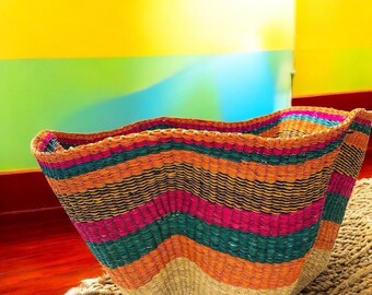 May include: A handwoven basket with a wavy rim, featuring horizontal stripes of orange, pink, green, and black. The basket sits on a textured, light brown surface, showcasing a blend of natural and vibrant colors.