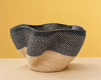 May include: A woven basket featuring a two-tone design with a natural tan base and a dark black upper section. The basket has a wide, undulating, wavy rim and a textured, hand-woven construction that creates a distinct, organic shape.