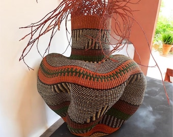 Könnte beinhalten: Ein großer, geflochtener Korb mit einer einzigartigen, wellenförmigen Form. Der Korb ist aus natürlichen Materialien gefertigt und hat ein braunes, orangefarbenes und grünes Farbschema. Der Korb ist mit einem geometrischen Muster verziert.