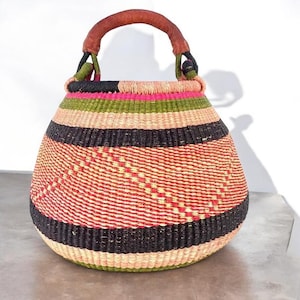 May include: A large, round, woven basket with a leather handle. The basket is made of natural fibers and has a colorful striped pattern in shades of pink, green, black, and beige.