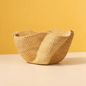 Peut inclure: Un panier tissé à la main de couleur beige clair, doté d'un bord large, ondulé et sinueux, ainsi que d'une base profonde et arrondie. Le panier est fabriqué en fibres naturelles avec un tissage vertical serré et visible.