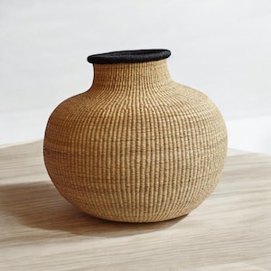 May include: A large, round, woven basket vase with a black rim. The vase is made of natural materials and has a light brown color.
