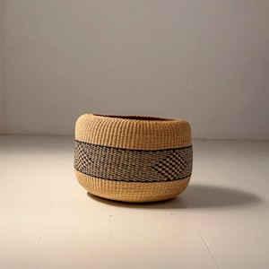 May include: A large, round, woven basket with a black and white geometric pattern. The basket is made of natural materials and has a light brown color.