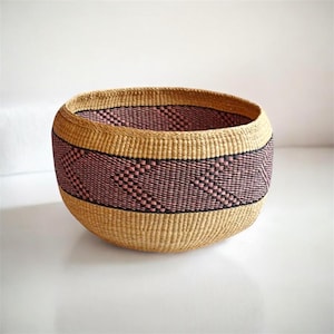 May include: A round woven basket with a natural tan color and a band of pink and black geometric pattern. The basket has a wide opening and a rounded base, suitable for storage or display.