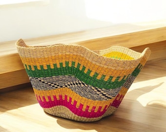 May include: A woven basket featuring a wavy, flared rim and a vibrant, multi-colored pattern. The design incorporates horizontal bands of natural tan, bright yellow, vivid green, deep black, and hot pink in a repeating geometric weave.