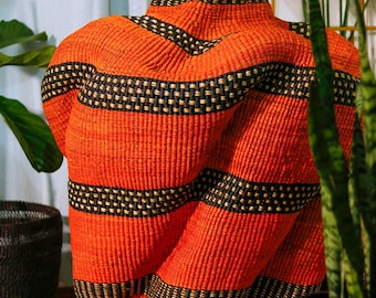 May include: A woven orange and black decorative basket with a tall, vase-like shape. The basket features horizontal black bands with a dotted pattern, and a black rim. The basket is a unique home decor item.
