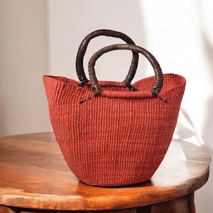 May include: A woven red basket with two dark brown leather handles. The basket has a rounded shape and is sitting on a wooden table. The handles are curved and attached to the top of the basket.