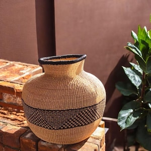 May include: A large, round, woven basket with a black rim and a black and white geometric pattern around the middle. The basket is made of natural fibers and has a rustic, earthy look.