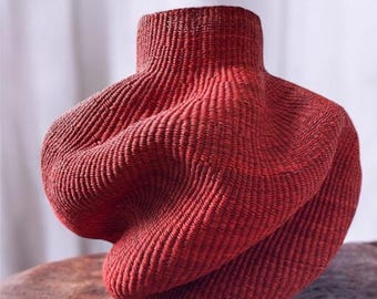 May include: A striking red woven vase with an unusual, swirling form. The vase features a cylindrical neck and a rounded body, displaying detailed texture and a deep, warm hue. This decorative item is ideal for adding an artistic element to any room.