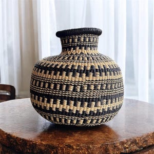 May include: A black and natural woven basket with a round body and a narrow neck. The basket has a geometric pattern.