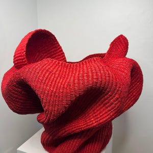 May include: A vibrant red woven basket with an organic, sculptural form. The basket has a wide opening and a textured surface, with folds and curves that create a dynamic shape. The basket is displayed on a white pedestal, showcasing its artistic design.