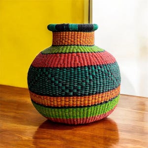May include: A large, round, woven basket with a wide mouth. The basket is made of natural fibers and is decorated with colorful stripes of red, green, orange, and black.