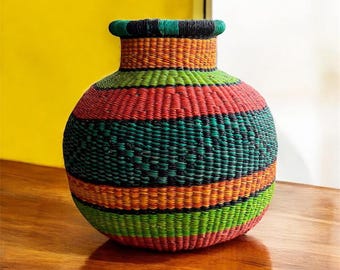 May include: A large, round, woven basket with a wide mouth. The basket is made of natural fibers and is decorated with colorful stripes of red, green, orange, and black.
